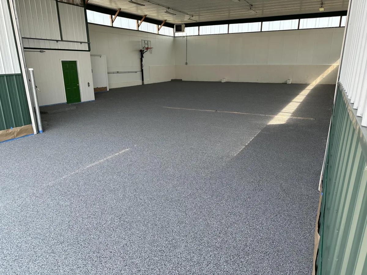 Spacious indoor sports court with speckled flooring, high ceiling, and basketball hoop. Walls feature green accents and high windows allow natural light.