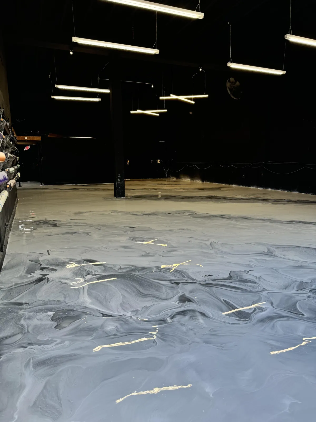A dimly lit industrial space with glossy, marbled flooring, overhead lights, and materials stacked on shelves to the left.