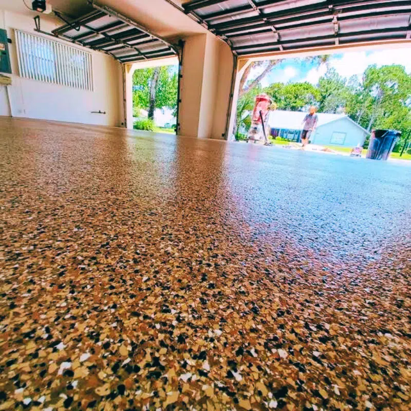 Shiny, textured garage floor in focus; open garage doors reveal two persons outside. Greenery and a house are visible in the background.