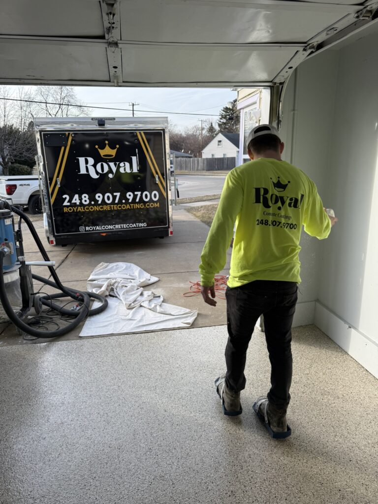 A person applies coating inside a garage. A truck with "Royal Concrete Coating" branding is visible outside, parked on the driveway.