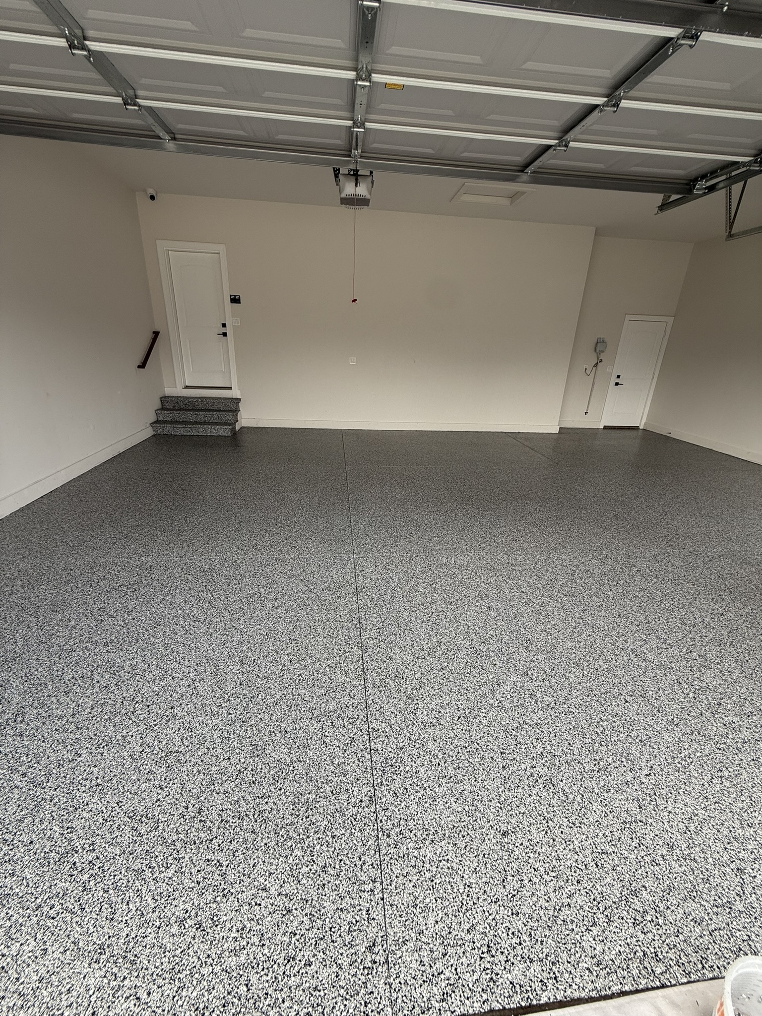 The image shows an empty garage with a speckled floor, white walls, closed doors, and a ceiling-mounted garage door opener.
