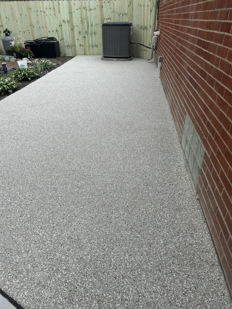 A newly coated concrete patio with a speckled finish, beside a red brick wall, bordered by plants and a wooden fence.